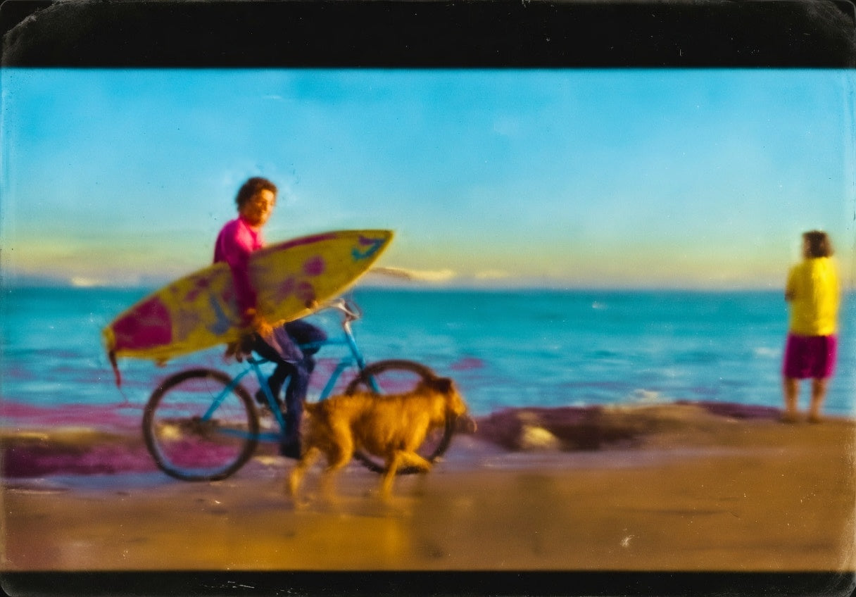 Shawn "Barney" Barron riding his bike along Steamer Lane on the Westside of Santa Cruz, carrying his surfboard, with his dog Foxy running alongside him.
